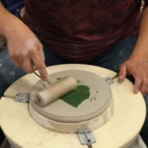 showing hands holding a roller that is pressing a leaf into wet clay